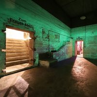 Innenraum eines Bunkers mit grün leuchtender Farbe an den Wänden, Türen mit Beschriftungen »Zum Frauen-Abort« und »Ausgang«