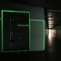 Dark corridor in the Pankstraße civil defence facility with fluorescent green markings on door frames and walls, door on the left labelled ‘AUSGANG’ (EXIT)
